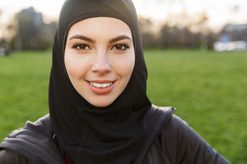 Portrait of young athletic muslim woman dressed in religious black hijab doing workout in green...