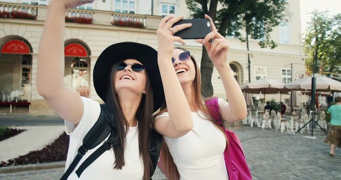 Two beautiful ladies dressed in casual clothing with backpacks taking photos of city architecture on smartphones, tourists enjoying trip