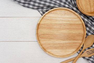 Empty round wooden plate with spoon, fork and grey gingham tablecloth on white wooden table. Top...