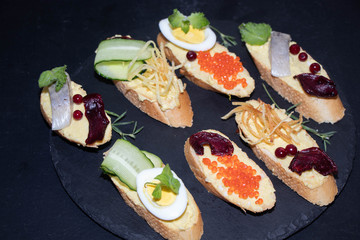 Baguette smeared with minced meat, on it pieces of herring, red onion, red caviar, all on a black, stone plate.