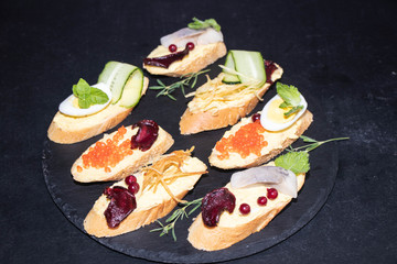 Baguette smeared with minced meat, on it pieces of herring, red onion, red caviar, all on a black, stone plate.