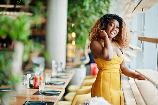 Glamour African American Woman In Yellow Dress Posed At Restaurant Near Windows.