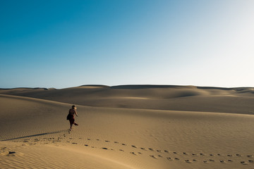 Fototapeta na wymiar evocative young confident woman walking on her own path on the desert sand with red dress in the middle of dunes on hot summer day with clear blue sky