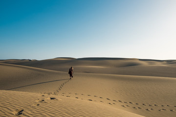 evocative young confident woman walking on her own path on the desert sand with red dress in the middlw of dunes on hot summer day with clear blue sky