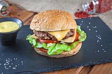 Fresh tasty burger with cheese, lettuce, tomato, cucumber on black stone with sauce. American fast food. Cheeseburger with copy space on wooden background. Close up, selective focus. Food. Grill menu
