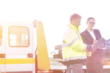 Businesswoman signing report while talking to tow truck driver against sky