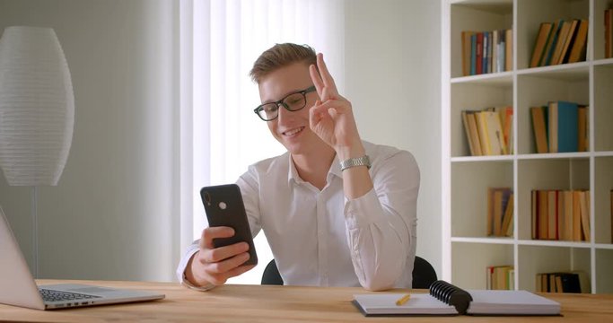 Closeup portrait of young handsome caucasian businessman in glasses taking selfies on the phone smiling cheerfully showing two fingers indoors in the office