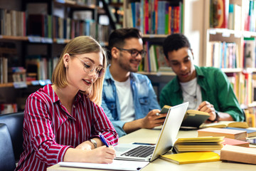 Three young students study in the school library, female student using laptop for researching online.