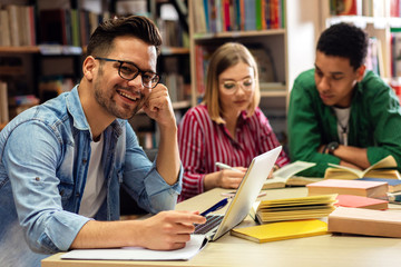 Three young students study in the school library, male student sitting in front of laptop and looking at camera.
