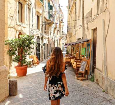 Fototapeta Woman in beautiful flowered dress walking in street of Syracuse, Sicily. Rear view of cheerful and happy girl in typical Italian old cozy street. Pretty sexy fashion model in Italy.
