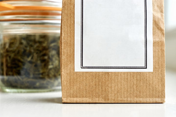 Fototapeta premium Brown kraft paper bag with white blank label in front of a bale glass jar and orange rubber gasket with dried herbs or tea on a white marble background. For use as a health template with copy space. 
