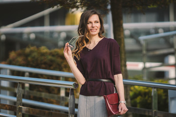 Caucasian adult lady woman in glasses posing near office building outside. Business lady in skirt holds hand glasses for vision bag in hand. Profession teacher assistant manager