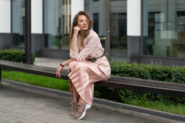 Fototapeta premium Young European in a jacket, pleated skirt, coral-colored sneakers resting on a bench in the street near the business center. Horizontal portrait of a beautiful girl.