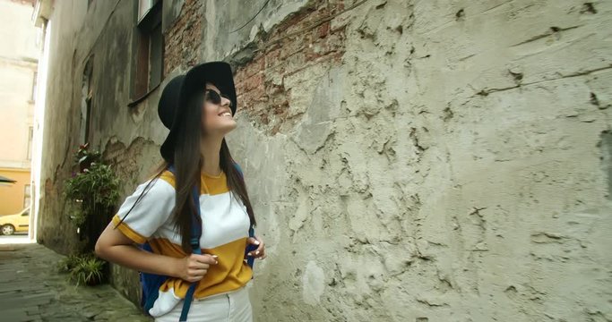 Beautiful female backpacker tourist in sunglasses and hat enjoying tour to ancient city, walking on narrow streets