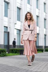 White girl walks down the street in a pink jacket, pleated skirt and sneakers. The territory of the business center. Great mood. Growth portrait.