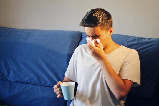 Cold man sitting on the couch with a handkerchief and a cup of tea.