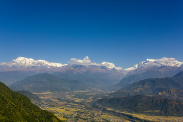 Naklejka premium bright green slope of a hill against the backdrop of the city in a mountain valley and the snowy ridge of Annapurna under a clear dark blue sky