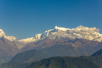 green forested slopes against the background of desert mountains and snowy peaks of Annapurn with white clouds under a clear blue sky