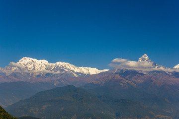 wooded hillsides in a mountain misty valley with snowy peaks of the Annapurna Ridge with white clouds under a clear blue sky