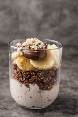 Dessert in a glass of yogurt, granola, banana and chocolate with cookies. Breakfast at the table on a gray background, copy space, close-up.