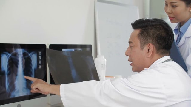 Medium shot of male Asian doctor sitting at desktop, comparing x-ray pic from computer screen with one in his hands and discussing them with his female coworker standing nearby