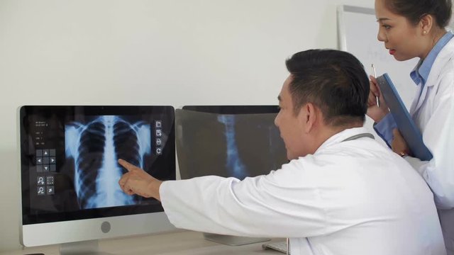 Rear view of male Asian doctor sitting at desk, looking at computer screen with x-ray pic, comparing it with one in his hands and discussing them with his female colleague standing nearby