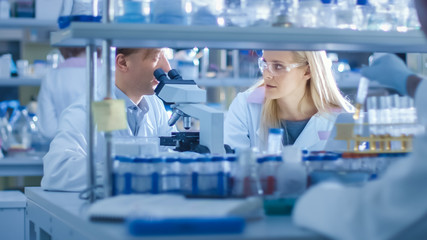 Team of a Male and Female Research Scientist working with a Microscope in a Laboratory. Having a Discussion about Genetics and Pharmaceutical Studies and Researches.