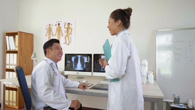 Lockdown of male Asian doctor sitting at desktop, looking at computer screens with x-ray images then coming his female colleague and starting conversation