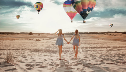 At sunset in the desert, two girls are walking along the sand against the background of balloons in...