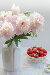 Obraz na płótnie Canvas Pink peonies in a white vase and a bowl of strawberries in the background