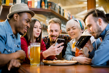 Young friends sharing pizza in a indoor cafe. Multiracial happy millennial friends having fun eating pizza together, laughing at funny joke and using smartphone