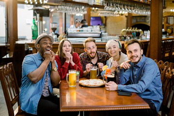 Cheerful multiracial friends having fun eating in pizzeria. Five friends emotionally watching sports on tv while eating pizza with drinks in cozy cafe