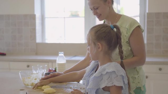 Side view of young woman and her little daughter cooking cakes at home in the kitchen. The girl finishing work and high five with mother. Mom and child spending time together. Real happy family.