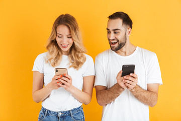 Portrait of curious young couple man and woman in basic t-shirts smiling while peeking at each other cellphones