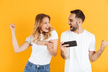Portrait of joyful young couple man and woman in basic t-shirts smiling and clenching fists while playing video games on cellphones