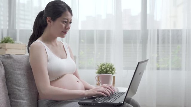 young asian pregnant woman with laptop computer typing keyboard. Beautiful expectant lady working on notebook pc while sitting on couch in living room. smiling future mom with cup of black coffee