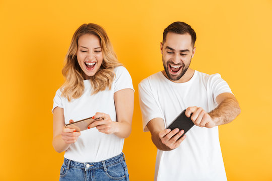 Portrait of excited young couple man and woman in basic t-shirts smiling and competing while playing video games on cellphones