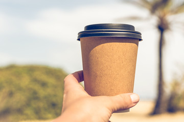 Woman's hand with paper cup of coffee on the palm trees background.