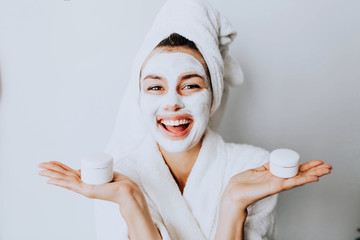 woman has fun with a facial mask.Young girl with facial mask, holding bottles of cream  looking at camera over white background. Cosmetic procedure. Beauty spa and cosmetology.Spa Facial Mask. Spa day