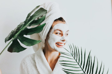 Young beautiful woman in face mask of therapeutic white mud and tropical leaves. Spa treatment,...