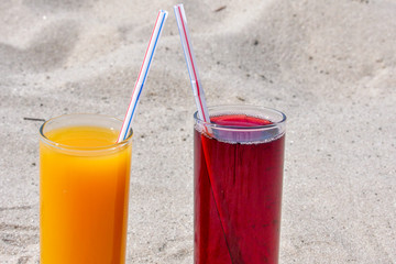 Two glass glasses with juice of mango and cherry with tubules. Sandy beach on a summer day