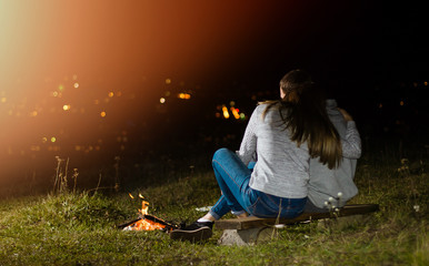 Happy couple in silhouette, sitting near a fire. Night sky with clouds. city lights on the background