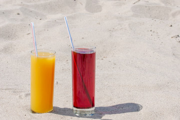Two glass glasses with juice of mango and cherry with tubules. Sandy beach on a summer day