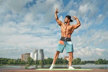 Fototapeta na wymiar Professional bodybuilder posing on lake pier with hands up.