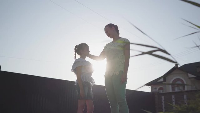 Cute mom and adorable little daughter standing on the street in the summer evening talking. Mom kisses a girl in the forehead. Relationship mom and daughter. Real happy family.