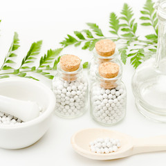 Fototapeta na wymiar glass bottles with small round pills near mortar and pestle, wooden spoon, jar and green leaves on white