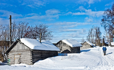 Nearly abandoned traditional fishers settlement on Kola Peninsula; old woman in blue jacket with trekking poles in hands passing by three grey wooden houses with roofs, covered with snow