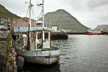 View on industrial harbour: Old, rusty, abandoned fishing boat moored in docks, big green hills,...