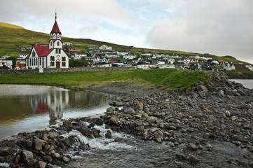 View on red & white church on lakeside, Faroe Islands; streamlet with small rocks and green grass forming coastline, small scandinavian town in the background;