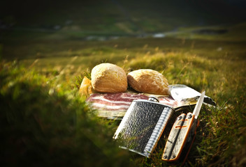 Appetizing picnic placed on green hills of Faroe Islands, Europe. Backed bread roll, fresh bacon, black & silver flask with whiskey, grey touristic folding multitool knife in front.